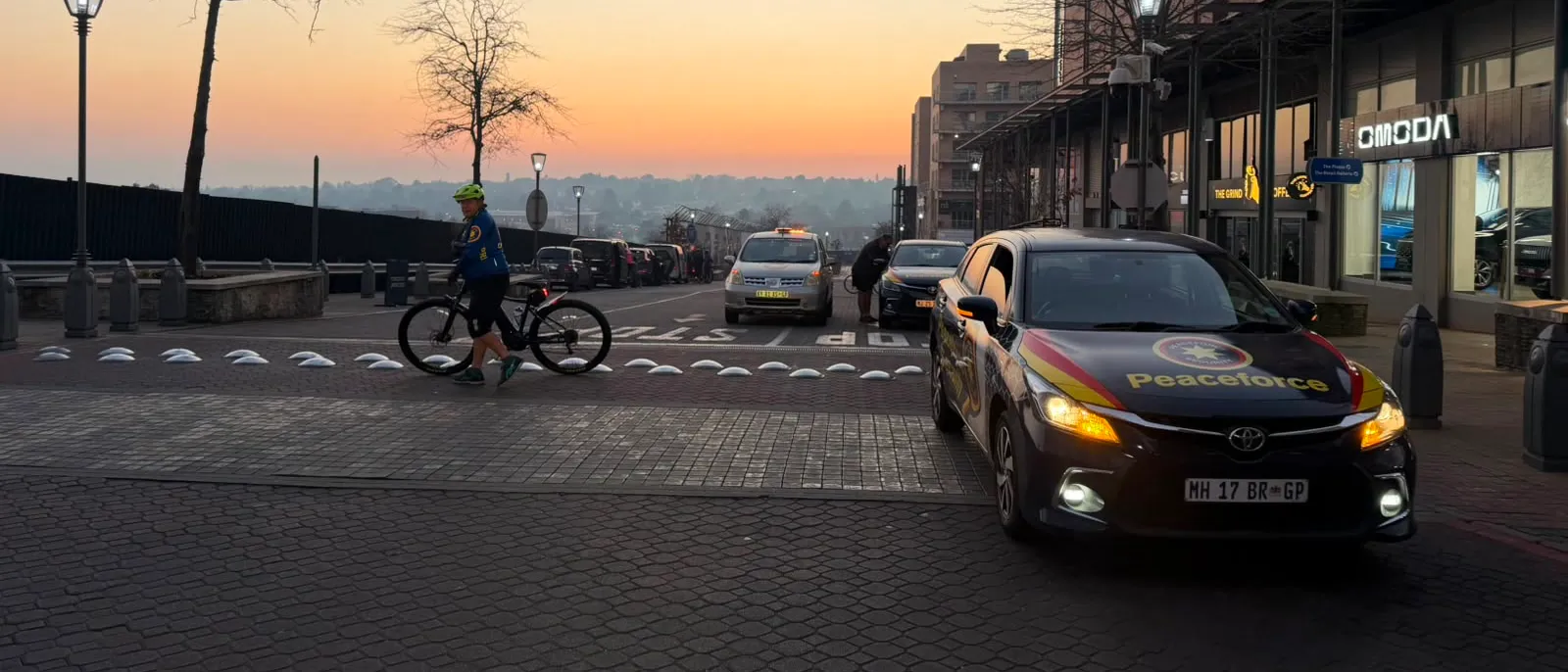 Sunrise over Melrose Arch as a rider wheels their bicycle to the start, with a Peaceforce follow car alongside
